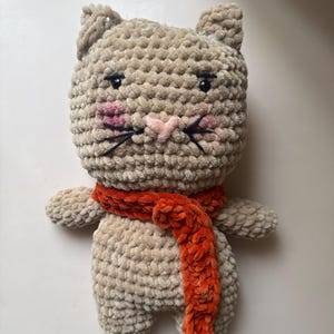 May include: A beige crocheted cat plush toy made from soft, textured yarn. The cat features embroidered black eyes, a small pink heart-shaped nose, black whiskers, and pink blush on its cheeks. It wears a bright orange crocheted scarf.