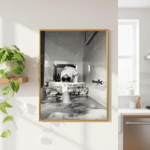 Puede incluir: Una impresión de arte mural enmarcada en blanco y negro que muestra a una persona en una bañera con una toalla en la cabeza y gafas de sol oscuras mientras lee un periódico titulado The Times. La imagen está en un marco de metal dorado.