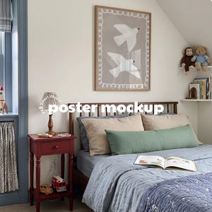 May include: A framed poster mockup featuring two white birds on a taupe background with a white looped border, displayed above a bed with blue and beige bedding, a red wooden nightstand, and children's toys in a bedroom setting.