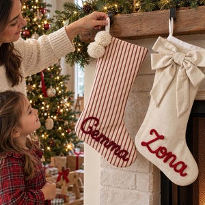 Peut inclure: Deux chaussettes de Noël personnalisées suspendues à une cheminée. L'une présente des rayures verticales rouges et blanches avec deux pompons blancs et le prénom Gemma brodé en rouge. L'autre est en tissu crème uni avec un grand nœud.