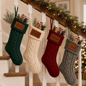 Puede incluir: Cuatro calcetines navideños de punto trenzado en verde oscuro, crema, rojo y gris. Cada calcetín tiene un parche rectangular de cuero marrón grabado con un nombre: Bruce, Pamela, James o Noah. Los calcetines están rellenos de ramas y bayas.