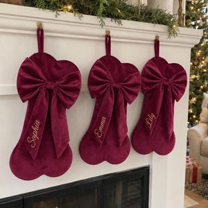 May include: Three deep burgundy velvet Christmas stockings shaped like dog bones, each featuring a large matching velvet bow at the top. The stockings are personalised with the embroidered names Sophia, Emma, and Lily in gold cursive script.