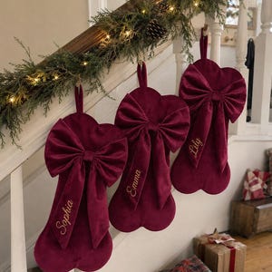May include: Three deep burgundy velvet Christmas stockings shaped like dog bones, each featuring a large decorative bow at the top. The stockings are personalised with gold embroidered names: Sophia, Emma, and Lily, hanging from a staircase railing.