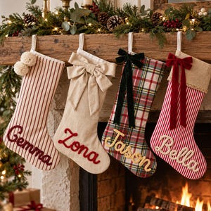 Peut inclure: Quatre chaussettes de Noël suspendues en rangée. La première est rayée rouge et blanc avec deux pompons et le nom Gemma. La deuxième est beige uni avec un grand nœud et le nom Zona. La troisième est à carreaux avec un nœud noir et le nom Jason. La quatrième est rayée rouge et blanc avec un revers en toile de jute, un nœud rouge et le nom Bella.