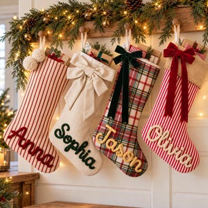 Peut inclure: Quatre chaussettes de Noël personnalisées suspendues en rangée. La première est rayée rouge et blanc avec le nom Anna. La deuxième est crème avec un nœud et le nom Sophia. La troisième est à carreaux avec un nœud vert et le nom Jason. La quatrième est rayée rouge et blanc avec un nœud rouge et le nom Olivia.