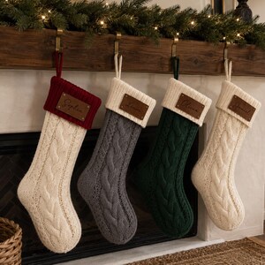 Puede incluir: Cuatro calcetines navideños de punto trenzado colgados en fila. El primero es crema con puño rojo, el segundo es gris con puño crema, el tercero es verde oscuro con puño crema y el cuarto es crema con puño crema. Cada uno tiene una etiqueta de cuero.