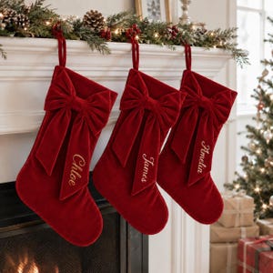 May include: Three deep red velvet Christmas stockings hanging in a row. Each stocking features a large, matching velvet bow at the top and is personalized with gold embroidered names: Chloe, James, and Amelia, stitched onto the long bow tail.