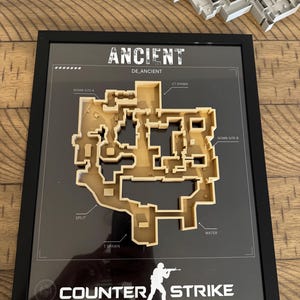 以下が含まれることがあります： Counter-StrikeのAncientレベルの3Dマップが額装されており、Bomb Site A、Bomb Site B、CT Spawn、T Spawn、Split、Waterのラベルが付いています。黒いフレームには、兵士のアイコンと共にANCIENT、DE_ANCIENT、COUNTER STRIKEの文字が表示されています。
