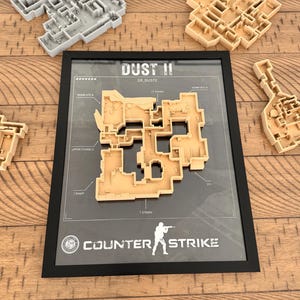 Könnte beinhalten: Ein gerahmtes Wandbild mit einer beigen 3D-Karte des Levels Dust II aus Counter-Strike, beschriftet mit T Spawn, T Ramp, Upper Tunnels, Bomb Site A und CT Spawn. Drei weitere unmontierte 3D-Kartenmodelle in Beige und Grau liegen daneben.
