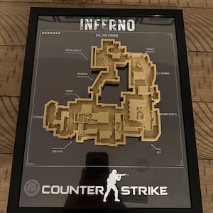 Peut inclure: Une carte 3D encadrée du niveau Inferno de Counter-Strike. La carte de couleur beige présente des étiquettes pour des lieux comme Bomb Site B, Banana, Ramp, T Spawn, Bridge, Pit, Bomb Site A, Truck et CT Spawn. Le cadre noir inclut le titre.