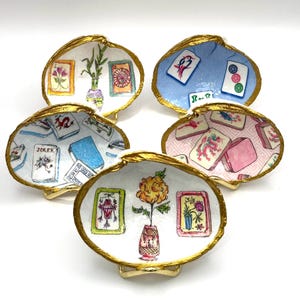 Op de afbeelding: Vijf decoratieve schelpschaaltjes met goudkleurige randen, elk voorzien van handgeschilderde illustraties. De ontwerpen bevatten mahjongtegels, bloemstukken en een jokerkart. De schelpen zijn in een groep gerangschikt en tonen diverse kleurrijke motieven.