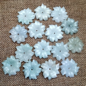 May include: A collection of jade flower pendants in various shades of green and white. Each pendant is carved in a floral design with detailed petals and a central hole. The pendants are arranged on a textured brown surface.