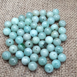May include: A collection of round, polished jade beads in varying shades of green and blue-green. Each bead has a hole drilled through the center, suggesting they are for stringing into jewelry. The beads are scattered on a textured, tan surface.