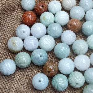 May include: A collection of jade green and brown round beads, some with a carved design. The beads are arranged on a brown burlap surface.