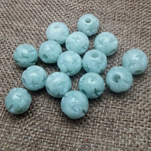May include: A collection of light green jade beads. Each bead is carved with a floral design and has a hole drilled through the center. The beads are round and have a smooth, polished surface. The beads are scattered on a brown, textured surface.