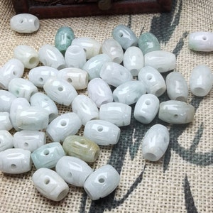 May include: A collection of oval-shaped, light green and white jade beads. Each bead has a hole drilled through the center. The beads are scattered on a textured, tan surface. The beads vary in color, with some having a more pronounced green hue.