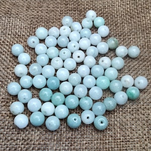 May include: A collection of light blue jade beads, each bead is round and smooth. The beads are arranged in a random pattern on a brown burlap surface.