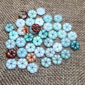 May include: A collection of jade flower-shaped beads in various shades of green, brown, and white. The beads are carved with intricate details and have a smooth, polished finish.