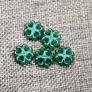 Puede incluir: Cinco botones verdes en forma de trébol de cuatro hojas. Los botones están hechos de un material translúcido y tienen un diseño ligeramente en relieve.