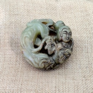 May include: A carved jade pendant featuring a figure with flowing robes and a serene expression. The light green stone has darker veining and is sculpted in a circular design. The pendant is likely for a necklace or other jewelry.