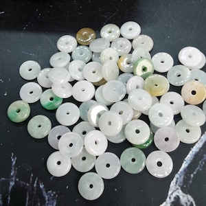 May include: A collection of jade donut-shaped beads in various shades of green, white, and tan. The beads have a smooth, polished surface and a hole in the center. These beads are suitable for jewelry making and crafting projects.