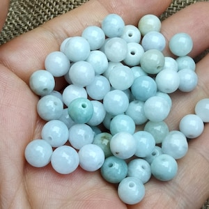 May include: A collection of light blue jade beads, each with a small hole drilled through the center. The beads are smooth and polished, and they are arranged in a loose pile.