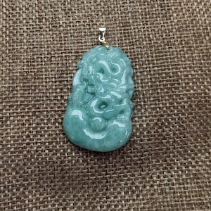 Puede incluir: Un colgante de jade verde tallado en forma de dragón. El colgante tiene una anilla de oro y está sobre un fondo de arpillera marrón.