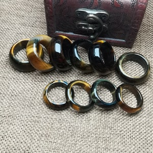 以下が含まれることがあります： 虎目石と黒瑪瑙で作られた10個のリングのコレクション。リングは積み重ねられ、茶色の布の表面に半円形に配置されています。