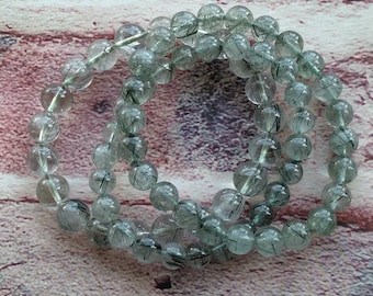 May include: Three clear bracelets with round beads. Each bead features dark green and black inclusions, creating a unique pattern. The bracelets are strung on a clear elastic cord, displayed on a light brown wooden surface.