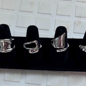 May include: Four silver-toned rings displayed on black velvet finger forms. The first ring is a wide band, the second is an open geometric shape, the third is a smooth asymmetrical piece, and the fourth features a ribbed, textured design.