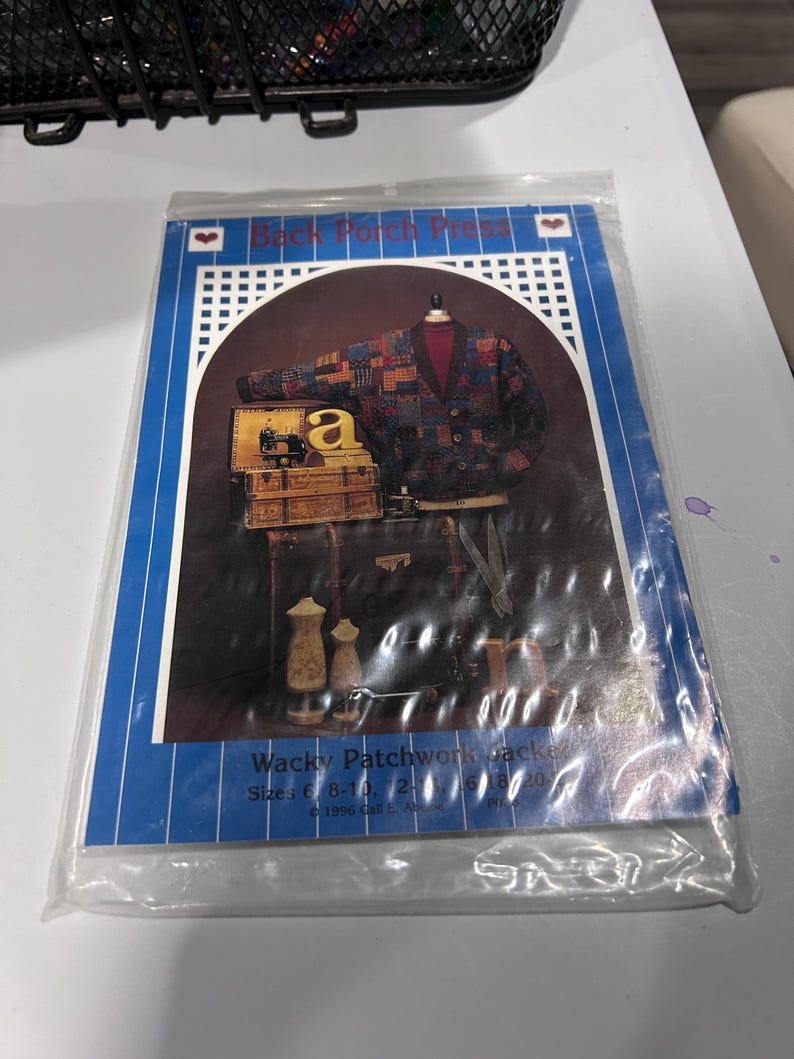 以下が含まれることがあります： 「Back Porch Press Wacky Patchwork Jacket」と書かれた裁縫パターンの封筒。表紙には、ダークカラーのパッチワークカーディガン、ミシン、2体のトルソー、ハサミが写っています。テキストにはサイズ6-20と1996年という記載があります。