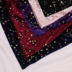 May include: A collection of velvet fabric swatches in black, deep red, purple, and light pink, each featuring a gold foil celestial print of scattered stars and constellations across the soft, textured surface of the material.