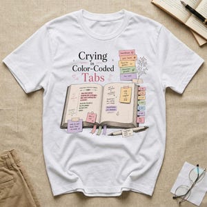 May include: A white short-sleeved t-shirt featuring a graphic of an open book with colorful sticky tabs and handwritten notes. The text reads Crying in Color-Coded Tabs. The design includes various pastel-colored sticky notes and a pen resting on the page.
