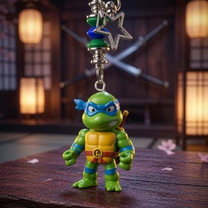May include: A green turtle action figure keychain featuring a blue mask, yellow chest plate, and brown belt with a letter L buckle. The figure is attached to a silver metal ring with a star-shaped charm and green and blue cylindrical beads.