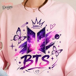 May include: A light pink crewneck sweatshirt featuring a central graphic of the BTS logo filled with a purple and pink galaxy nebula pattern. The design includes a crown, two butterflies, a planet, stars, hearts, and the text BTS in purple.