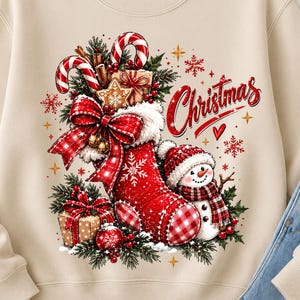 Può includere: Una felpa beige a girocollo con una stampa festiva raffigurante una calza natalizia rossa riempita di bastoncini di zucchero, un biscotto a forma di stella e regali. Un piccolo pupazzo di neve con cappello e sciarpa a quadri rossi.