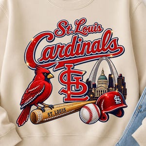 May include: A beige crewneck sweatshirt featuring a graphic of a red cardinal bird perched on a wooden baseball bat, a baseball, a red batting helmet, the St. Louis Gateway Arch, and the text St. Louis Cardinals in red and blue lettering.