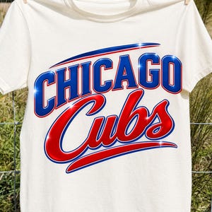 Puede incluir: Una camiseta de manga corta de color crema con el texto CHICAGO CUBS en un gráfico brillante y llamativo. La palabra CHICAGO está impresa en azul con un borde blanco, mientras que la palabra Cubs está en rojo con un borde blanco.