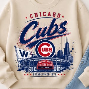 Puede incluir: Una sudadera de cuello redondo color beige con un estampado gráfico de los Chicago Cubs. El diseño incluye el nombre del equipo, el logotipo de los Cubs, una representación de Wrigley Field, una bandera W, el horizonte de Chicago y el texto Wrigley Field Home of Chicago Cubs Est. 1876.
