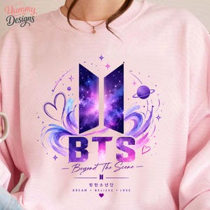 May include: A light pink crewneck sweatshirt featuring a graphic print of the BTS logo filled with a purple and blue galaxy pattern. The design includes the text BTS, Beyond The Scene, and Korean characters, surrounded by stars, planets, and hearts.