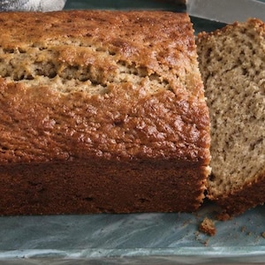 May include: A golden-brown loaf of banana bread with a cracked top crust, featuring two thick slices cut from the loaf. A silver knife rests beside the bread, which is displayed on a rectangular, dark teal, marbled-finish serving tray.