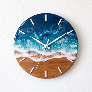 May include: A round wall clock featuring a resin art design that mimics an ocean shoreline. The bottom half is natural wood grain, while the top half displays swirling shades of deep blue and turquoise with white foam waves and white hands.