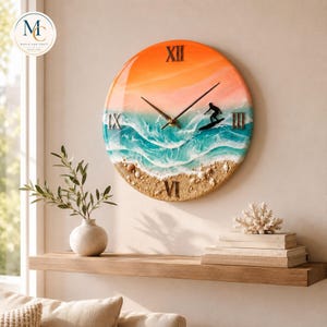 May include: A round wall clock featuring a resin ocean scene with a gradient orange sky, turquoise waves, and a sandy beach base with small shells. A black silhouette of a surfer on a board is visible. Roman numerals mark the hours XII, III, VI, and IX.