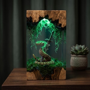 Puede incluir: Una lámpara rectangular de resina con un marco de madera natural de bordes irregulares. Dentro de la resina transparente, una serpiente de cascabel enroscada rodea una espada metálica, rodeada de musgo verde e iluminada por una luz verde.