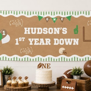 May include: A rectangular banner with a tan background and green striped borders featuring the text HUDSON'S 1ST YEAR DOWN. The design includes football graphics, a goalpost, balloons, and pennants in shades of green, brown, and white.