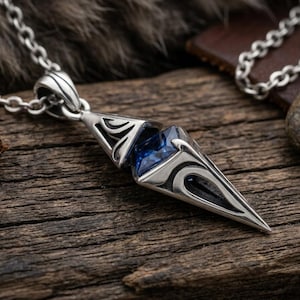 Peut inclure: Un collier pendentif en argent doté d'un boîtier métallique géométrique et pointu avec des motifs découpés complexes. Une pierre précieuse facettée bleu profond est sertie au centre du pendentif, suspendu à une chaîne à maillons en argent.