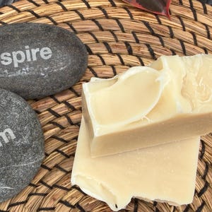 May include: Two smooth, dark gray oval stones with the words "inspire" and "dream" printed in white, accompanied by two rectangular bars of off-white, solid soap with a smooth, slightly textured surface and rounded edges on one side.