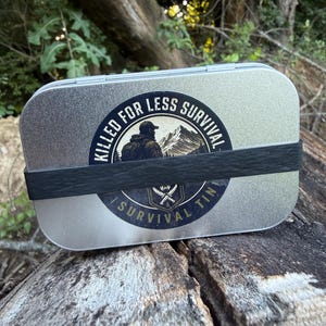 May include: A rectangular silver metal tin secured with a black elastic band. A circular sticker on the front features a mountain landscape, a person, and the text KILLED FOR LESS SURVIVAL and SURVIVAL TIN with a crossed knife graphic.