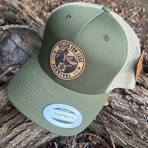 May include: An olive green trucker hat with a beige mesh back and a circular tan leather patch on the front. The patch features a black graphic of a person hiking in mountains with the text KILLED FOR LESS SURVIVAL and a Yupoong sticker.