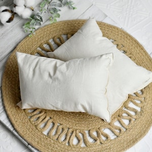 Puede incluir: Dos almohadas rectangulares blanquecinas descansan sobre una alfombra redonda de yute con un borde decorativo. Las almohadas están hechas de una tela texturizada. Ramas de algodón y vegetación en el fondo. Las almohadas están sobre una superficie de madera blanca.